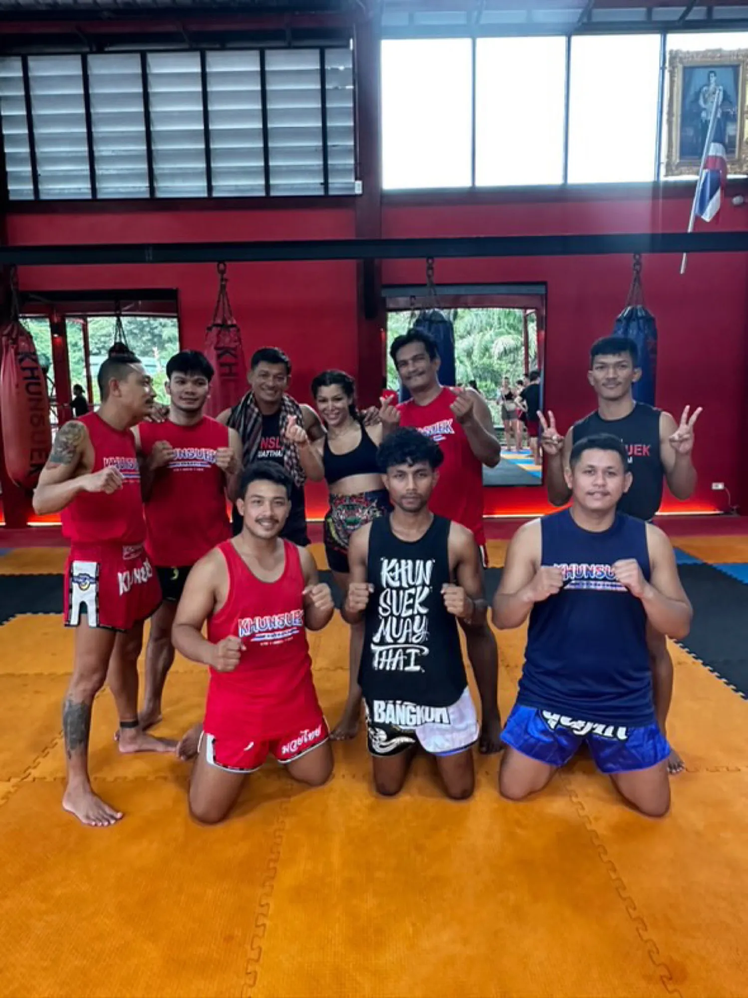 Photo de groupe dans la salle de Muay Thai : Sarah entourée de ses coachs, sacs de frappe et tatami derrière